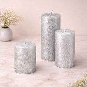 May include: Three silver pillar candles with textured surfaces and white wicks. The candles are of different heights and are set on a light pink surface. A small vase of white flowers is in the background.