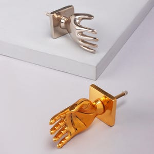 May include: Two hand-shaped cabinet knobs. One is silver, the other gold. Each has a square base and a screw for mounting. The hands are in a grasping pose, adding a unique touch to furniture. Suitable for cm measurements.