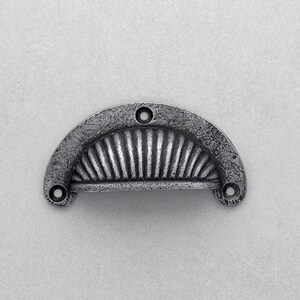 Puede incluir: Un tirador de cajón de metal plateado envejecido, con forma de media luna. El tirador presenta una superficie texturizada y un diseño en forma de abanico con ranuras verticales. Tiene tres orificios para el montaje.