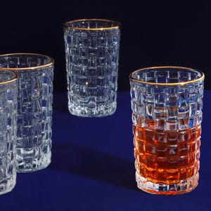 May include: Four clear drinking glasses with a textured, woven pattern. Each glass has a gold rim. One glass is filled with a reddish-brown liquid. The glasses are arranged on a dark blue surface.