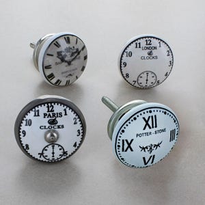 May include: Four cabinet knobs with clock face designs. The knobs have white faces with black numerals and text, including 'London' and 'Paris'. Each knob has a silver metal base and screw, suitable for furniture.