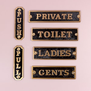 May include: A collection of black and gold metal signs with various text. The signs include "PUSH", "PULL", "PRIVATE", "TOILET", "LADIES", and "GENTS". The signs have a textured surface and are mounted with gold-colored screws.