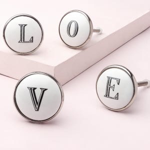 Peut inclure: Quatre boutons de meuble en céramique blanche épelant "LOVE". Chaque bouton a un bord en métal argenté et une lettre noire. Les boutons sont sur une surface rose clair.