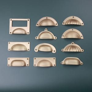 Könnte beinhalten: Eine Sammlung von elf matten, goldfarbenen Metall-Schubladengriffen und Muschelgriffen in verschiedenen Formen, darunter rechteckige Etikettenhalter und halbkreisförmige Griffe, jeweils mit vorgebohrten Schraublöchern zur Montage an Möbeln.