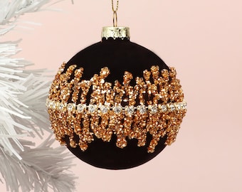 Puede incluir: Adorno navideño negro con detalles de cuentas doradas y una banda de brillantes transparentes. El adorno cuelga de un hilo dorado sobre un fondo rosa claro.