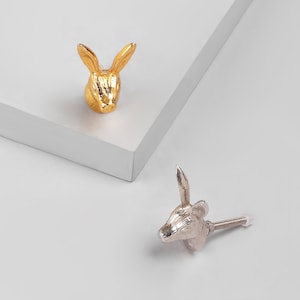 Puede incluir: Dos tiradores de cajón con forma de cabeza de conejo, uno dorado y otro plateado. La cabeza de conejo dorada está mirando hacia adelante, mientras que la cabeza de conejo plateada está mirando ligeramente hacia la derecha. Ambas cabezas de conejo tienen un tornillo en la parte posterior.