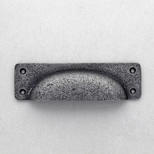Pode incluir: Um puxador de gaveta retangular e envelhecido, com uma pega em forma de concha. O hardware é cinza manchado com quatro orifícios para parafusos. O design evoca estilos vintage ou industriais. Ideal para móveis.