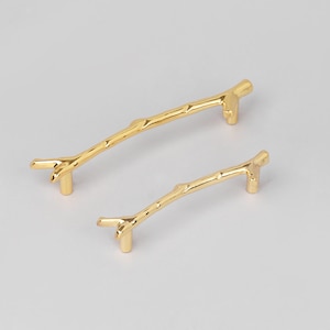 May include: Two gold-colored cabinet pulls in a twig design. The pulls are made of metal and have a textured surface, resembling natural branches. One pull is larger than the other.