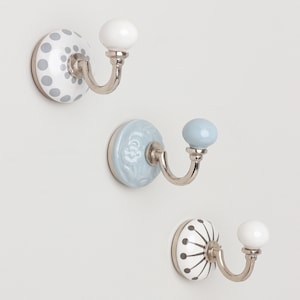 May include: Three decorative wall hooks with ceramic bases and metal hooks. The bases feature white, light blue, and patterned designs with grey and white accents. Each hook has a rounded knob at the end.