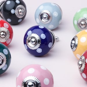 Könnte beinhalten: Eine Sammlung farbenfroher Keramik-Schrankknöpfe. Jeder Knopf hat eine andere Farbe, darunter Schwarz, Rot, Blau, Grün, Gelb und Rosa, und ist mit weißen Tupfen verziert. Die Knöpfe haben einen silbernen Metallsockel.