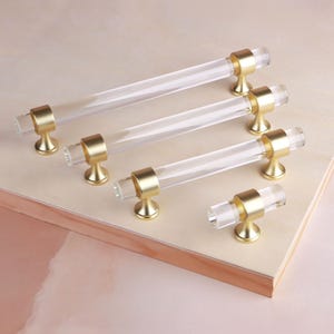 Könnte beinhalten: Ein Set aus vier Schrankgriffen in verschiedenen Längen, bestehend aus einer zylindrischen Stange aus klarem Acryl, die von zwei Pfosten aus gebürstetem Goldmetall gehalten wird. Das Set enthält einen langen Griff, zwei mittlere und einen Knopf.
