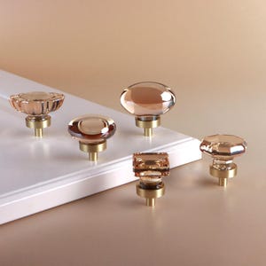 May include: Five cabinet knobs featuring translucent, peach-colored glass tops in various geometric shapes, including round, oval, and square, each mounted on a cylindrical brushed brass base, displayed on a white panel surface.