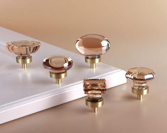May include: Five cabinet knobs featuring translucent, peach-colored glass tops in various geometric shapes, including round, oval, and square, each mounted on a cylindrical brushed brass base, displayed on a white panel surface.
