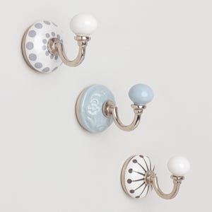 May include: Three decorative wall hooks with ceramic knobs. One hook has a white knob and a white base with grey polka dots. Another has a light blue knob and base. The third hook has a white knob and a white base with a black and white design.