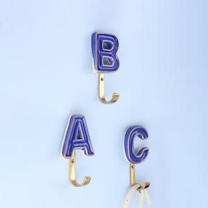 Peut inclure: Trois patères murales en forme de lettres A, B et C. Les crochets sont bleus avec un bord blanc et des crochets dorés. Ils sont montés sur un mur bleu clair. Une ceinture blanche est accrochée au crochet C.