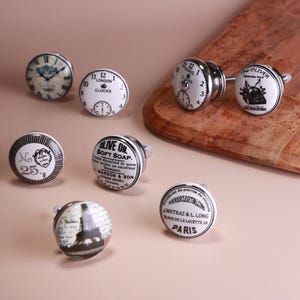 May include: A selection of decorative cabinet knobs with diverse designs. Some knobs feature clock faces, while others display text like "LONDON" and "PARIS". The knobs are white with black detailing and silver hardware.