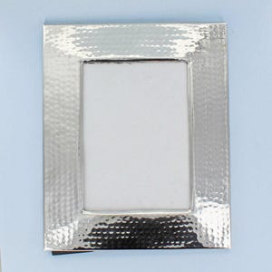 May include: A rectangular silver picture frame with a hammered texture. The frame has a simple, elegant design with a white interior space for a photograph or artwork. The frame is set against a light blue background.
