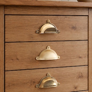 May include: A wooden chest of drawers featuring three horizontal drawers, each fitted with a polished brass cup pull handle. The handles are semi-circular in shape and are securely fastened to the wood grain surface with visible metal screws.
