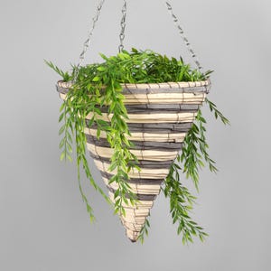 Op de afbeelding: Een hangende plantenbak met een conische vorm, gemaakt van geweven materiaal in afwisselende grijze en beige banden. De bak is gevuld met groene, bladrijke planten en wordt opgehangen aan een zilveren ketting.