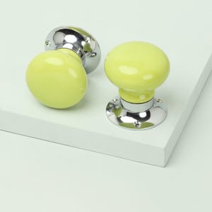 May include: Two yellow-green ceramic door knobs with a glossy finish. Each knob has a rounded shape and sits atop a shiny, chrome-colored base with screw holes. The knobs are displayed on a white surface.