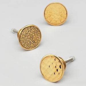 Może przedstawiać: Trzy złote gałki meblowe. Dwie gałki mają młotkowane wykończenie, a jedna gałka ma kwiatowy wzór.