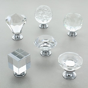 May include: Six clear crystal cabinet knobs with silver bases are displayed. The knobs showcase diverse geometric designs: a diamond, a bubble-filled sphere, a faceted sphere, a faceted dome, a cube, and a flat disc. These are ideal for furniture.
