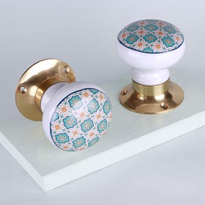 May include: Two white ceramic door knobs with a decorative teal and orange geometric pattern. The knobs have brass bases and are designed for easy installation. These decorative knobs add a touch of elegance to any door or cabinet.