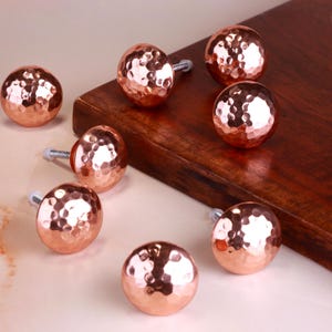 Könnte beinhalten: Acht kupferfarbene Schrankknöpfe mit gehämmertem Finish. Jeder Knopf ist kugelförmig mit einer strukturierten Oberfläche und einer silberfarbenen Schraube. Die Knöpfe sind auf einer hellen Oberfläche und einem Holzbrett angeordnet.