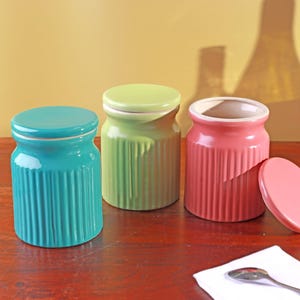 Puede incluir: Tres botes de cerámica en turquesa, verde y rosa, cada uno con una tapa a juego. Los botes tienen un diseño acanalado y están dispuestos sobre una superficie de madera. Una tapa rosa está a un lado. El fondo es amarillo cálido.