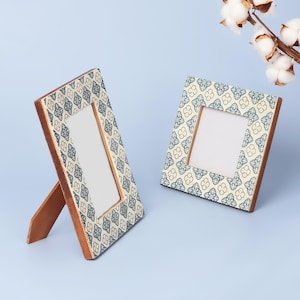 Op de afbeelding: Twee houten fotolijsten met een patroon in blauw en crème. De lijsten hebben een rechthoekige opening voor foto's en worden ondersteund door een houten ezelrug. Een takje katoen voegt een decoratief tintje toe.