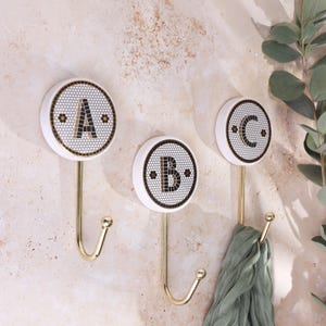 May include: Three wall hooks featuring circular white bases with black and gold mosaic tile patterns. Each hook displays a large letter A, B, or C in the center, constructed from small dark tiles. The hooks have a polished gold metal finish.