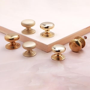 May include: A collection of six round cabinet knobs in gold-toned metal. Three knobs feature a polished, reflective finish, while the other three have a matte, satin finish. Each knob has a circular base and a smooth, rounded top surface.