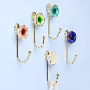Pode incluir: Cinco ganchos de parede decorativos com braços de metal dourado e botões de cerâmica coloridos em forma de gema, em tons de rosa, verde, azul e branco. Montados numa parede azul clara.