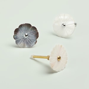 Peut inclure: Trois boutons décoratifs pour armoire avec un motif floral. Les boutons sont en nacre blanche et irisée. Deux boutons sont plats et un bouton a une vis dorée.