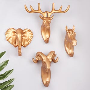 Puede incluir: Ganchos de pared dorados con forma de ciervo, elefante, caballo y carnero. Cada gancho está diseñado con detalles de animales y un gancho para colgar objetos. Los ganchos están montados en una pared blanca.
