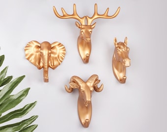 Peut inclure: Crochets muraux dorés en forme de cerf, d'éléphant, de cheval et de bélier. Chaque crochet présente des détails animaliers et un crochet pour suspendre des objets. Les crochets sont montés sur un mur blanc.