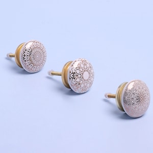 May include: Three white ceramic cabinet knobs with gold geometric designs. The knobs have a brass screw attachment.