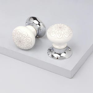 May include: Two white ceramic door knobs with a floral design in a light gray color. The knobs have a rounded shape and are mounted on shiny chrome bases. The knobs are on a white surface.