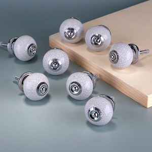 May include: A collection of eight white ceramic cabinet knobs with a crackle glaze pattern. Each knob has a silver-toned metal base and screw. The knobs are arranged on a light blue surface and a wooden block.