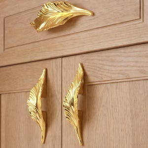 May include: Gold-coloured leaf-shaped cabinet hardware. The image shows a single, large leaf-shaped handle above two matching handles. The hardware is mounted on a light-coloured wooden cabinet door. The handles have a detailed, textured design.