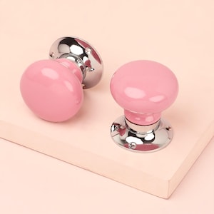 May include: Two pink ceramic door knobs with a glossy finish. The knobs have a rounded shape and are mounted on a shiny, silver-colored base. The door knobs are set against a light pink background.