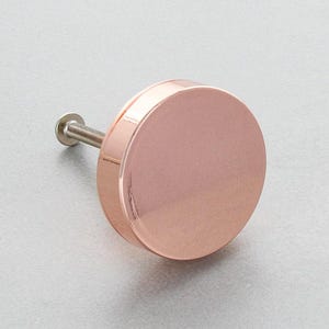 May include: A rose gold cabinet knob with a circular design. The knob has a polished, reflective surface and a threaded screw for installation. Suitable for drawers, cabinets, and other furniture.