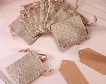 May include: A collection of small, beige burlap drawstring bags, some with twine closures. Two brown paper tags with scalloped edges are also visible. One bag contains a wrapped candy. The items are arranged on a light pink surface.