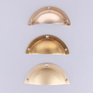 Pode incluir: Três puxadores de gaveta semicirculares dispostos verticalmente. O puxador superior tem um acabamento dourado polido, o do meio apresenta uma textura de latão escovado e o inferior tem um acabamento dourado mate e liso.