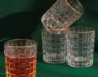 May include: Four clear drinking glasses with a textured, woven pattern. One glass is filled with a reddish-brown liquid. Each glass has a gold rim. The glasses are arranged on a dark green surface.