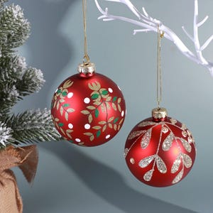Può includere: Due palline di Natale rosse con disegni di foglie dorate e verdi e pois bianchi. Una pallina ha un motivo a foglie argentate glitterate. Entrambe sono appese a fili d'oro, perfette per le decorazioni natalizie.