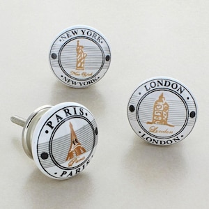 May include: Three white ceramic cabinet knobs with city-themed designs. One features the Statue of Liberty with "New York" text, another the Eiffel Tower with "Paris" text, and the third the Houses of Parliament with "London" text.
