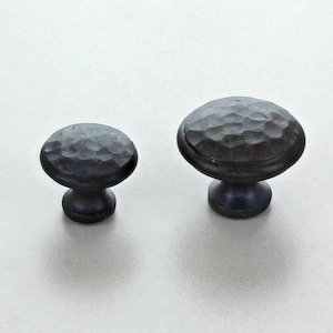 Puede incluir: Dos tiradores de armario de bronce oscuro con textura martillada. Los tiradores tienen una parte superior redondeada y una base corta y cilíndrica. Los tiradores se muestran sobre una superficie clara.
