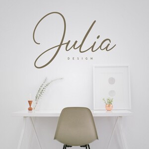 May include: A white desk with a white chair, a white picture frame, a vase with dried branches, and a small wooden candle holder. The wall behind the desk has the text "Julia Design" written in brown cursive.