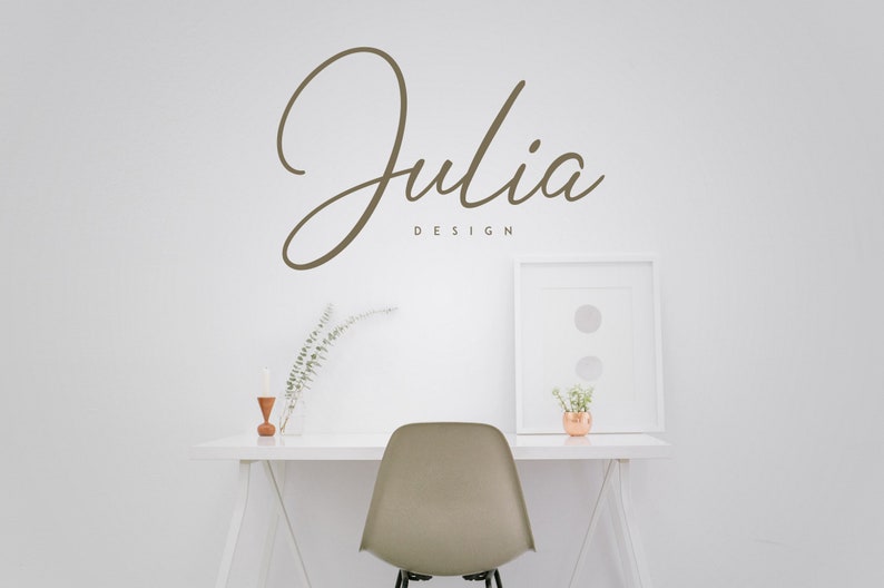 May include: A white desk with a white chair, a white picture frame, a vase with dried branches, and a small wooden candle holder. The wall behind the desk has the text "Julia Design" written in brown cursive.